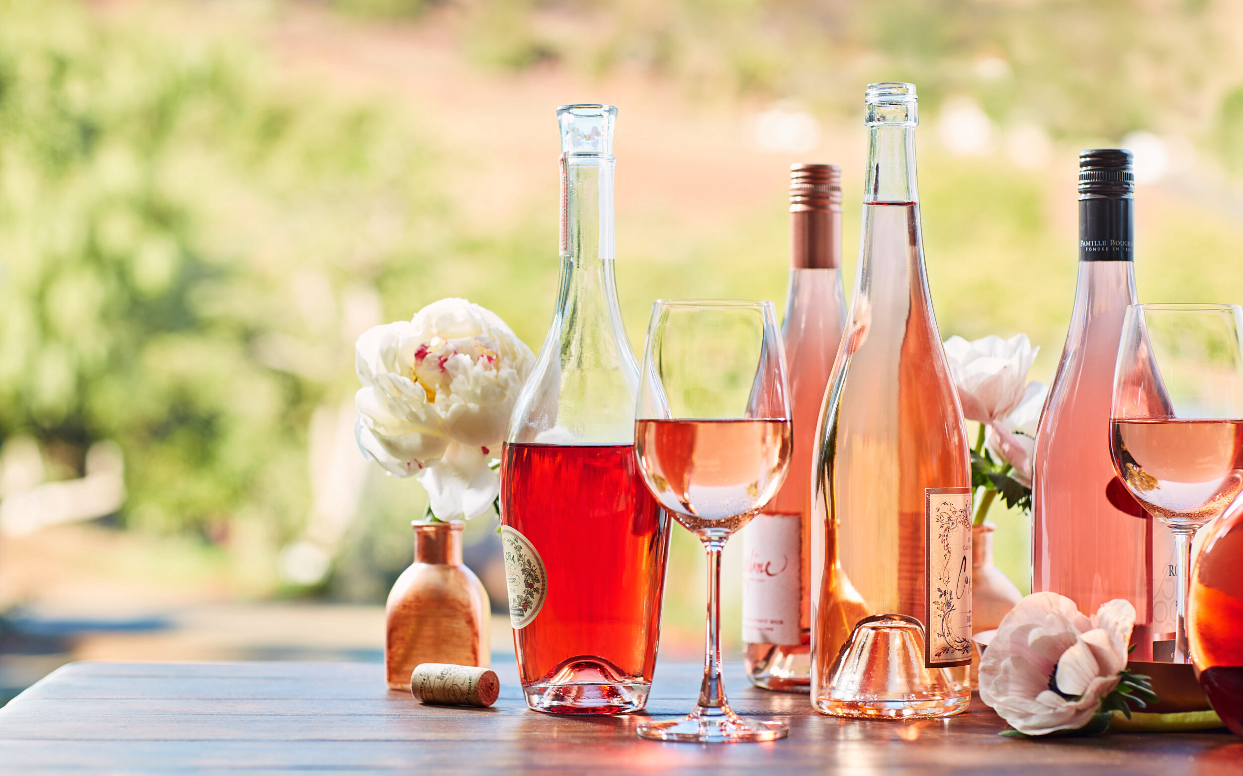 Four bottles of Rose Wine and two glasses of rose wine are set on a table outdoors with a vineyard out of focus in the background.  Image is taken by photographer Vinnie Finn who is repped by Andrea Stern of SternRep